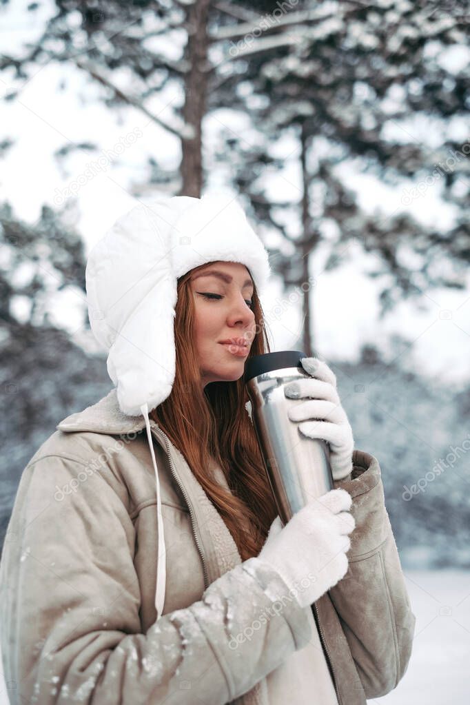 beautiful young girl in a white fur hat in the winter forest drinks tea from a thermos. woman drinks tea from a thermo mug. portrait of a beautiful girl in winter