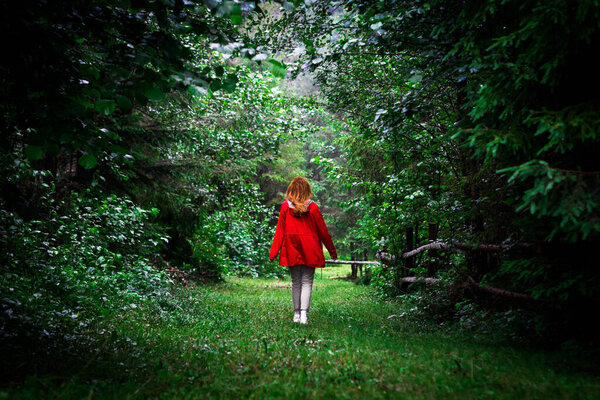 A young girl in a red jacket walks through a green forest in the mountains alone on self-isolation. woman spends time alone with nature. rest in the forest, meditation in nature