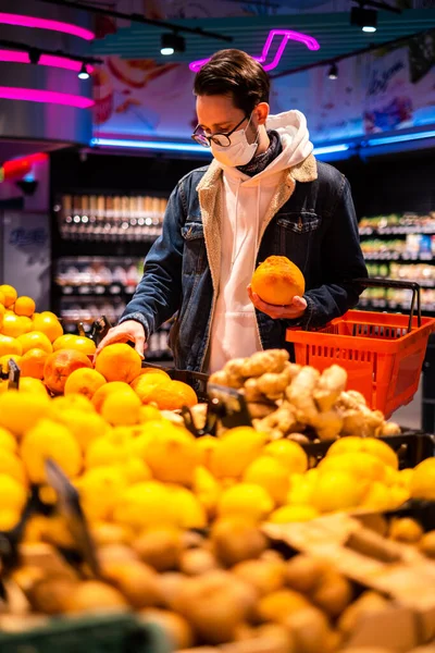 a man in a protective mask in a supermarket chooses fruits and citrus fruits. man during a pandemic buys groceries in the store. courier picks up an order in a supermarket