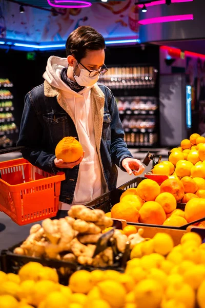 a man in a protective mask in a supermarket chooses fruits and citrus fruits. man during a pandemic buys groceries in the store. courier picks up an order in a supermarket