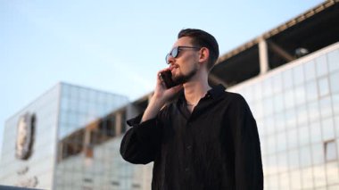 Güneş gözlüklü, siyah gömlekli genç ve yakışıklı bir adam iş merkezinin önünde telefonla konuşuyor.