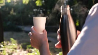 Bir kadın termostan bir fincana bitki çayı döküyor. Güneşli bir yaz sabahının erken saatlerinde, ellerinin kapalı olduğu bir yerde.