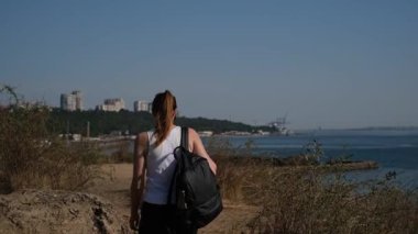 Sıcak ve güneşli bir günde sırt çantasıyla denizin yukarısında yürüyüş yapan şortlu genç bir kadın.