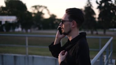 Güneş gözlüklü, siyah gömlekli genç ve yakışıklı bir adam iş merkezinin önünde telefonla konuşuyor.