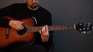 Yakışıklı bir esmer adam karanlık bir stüdyoda sandalyede otururken gitar çalıyor.