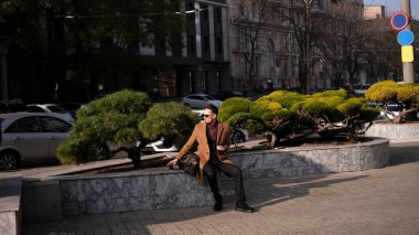 Gözlüklü ve ceketli genç adam güneşli bahar gününde iş merkezinin önünde kahve içiyor.