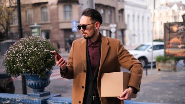 Bir paket taşıyan şık bir işadamı ve şehir caddesinde akıllı telefon kullanıyor.