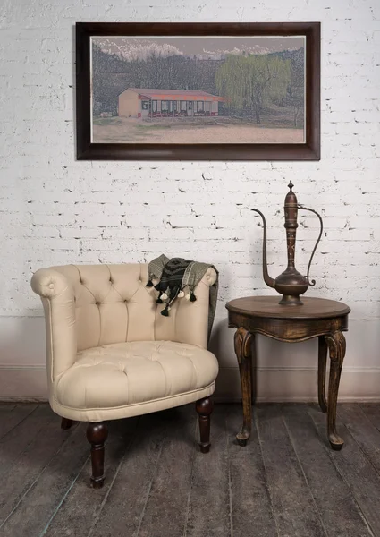 Oude beige fauteuil, messing theepot, ingelijste schilderijen en antieke tafel — Stockfoto