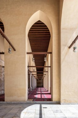 Grand interior of the Mosque of al-Zahir Baybars, Cairo, Egypt. Arches, columns, and prayer rugs with dramatic sunlight and shadows, highlighting Mamluk architecture.
