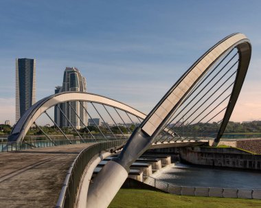 Lakeside pedestrian bridge at Taman Seri Empangan, Putrajaya, Malaysia. Modern arched design over cascading waterfalls, with contemporary skyscrapers visible under a clear blue sky.