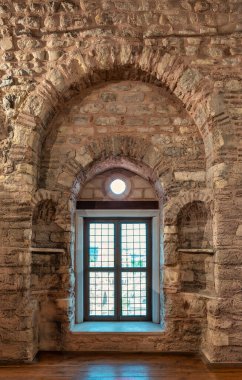 Textured stone and brick wall interior with an arched, grid-patterned window, circular transom, and side niches, inside a historic building