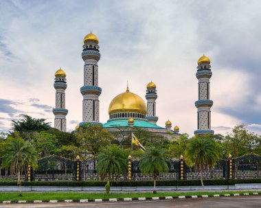 Jame Asr Hassanil Bolkiah Camii, Bandar Seri Begawan, Brunei. Altın kubbeler, minareler ve Brunei bayrağı bulutlu bir gökyüzünün altında yeşil yaprakların arasında görünür.