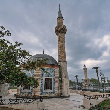 Hisar Camii ve onun karmaşık fayanslı minaresi ve arka plandaki İzmir Saat Kulesi bulutlu bir gökyüzünün altında. Konak Meydanı, İzmir, Türkiye.