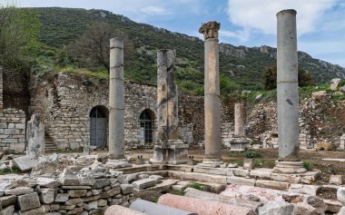 Efes 'in antik Roma kalıntıları, Selcuk, Türkiye. Görkemli yıpranmış sütunlar ve taş yapılar parçalı bulutlu bir gökyüzünün altındaki yemyeşil yamaçta duruyor..