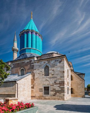 Mevlana Müzesi ve Anıtmezar, Konya, Türkiye. Kendine özgü turkuaz kubbesi ve ince bulutlu mavi gökyüzünün altında minaresi olan tarihi bir bina. Önplanda kırmızı çiçekler