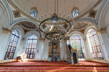 Aziziye Mosque interior, Konya, Turkey. Features a grand chandelier, red carpet, mihrab, minbar, and ornate dome with calligraphy. Sunlight fills the space through tall arched windows