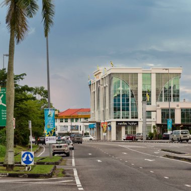 Bandar Seri Begawan, Brunei - 23 Şubat 2025: Modern mimari ve palmiye ağaçları bulutlu bir gökyüzü altında arabalarla hareketli bir sokak sahnesi oluşturuyorlar. Brunei bayrakları önde gelen bir binanın tepesinde dalgalanıyor