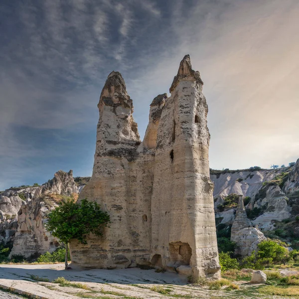 Goreme Açık Hava Müzesi, Goreme, Kapadokya, Türkiye. Antik kaya kesimli evleri ve parçalı bulutlu bir gökyüzünün altında yalnız bir ağacı olan gökdelenler. UNESCO Dünya Mirası
