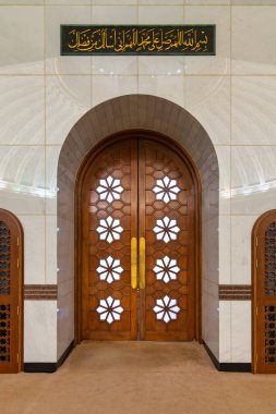 Jame Asr Hassanil Bolkiah Camii 'nin içi, Bandar Seri Begawan, Brunei Darussalam, geometrik yıldız desenleri ve Arapça kaligrafi plağıyla süslü ahşap bir kapı gösteriyor.