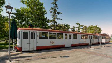Konya, Türkiye - 7 Haziran 2025: Arka planda yeşil ağaçlar bulunan güneşli Kulturpark Parkı 'ndaki kaldırımlı bir alanda, beyaz ve kırmızı renkli, eşsiz bir tramvay şeklinde büfe büfesi