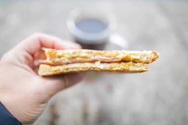 Bir fincan bulanık kahvenin önünde peynirli ve jambonlu sandviç tutarken.