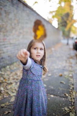 Kaldırımdaki küçük kız kameranın önünde bir sonbahar yaprağı tutuyor.