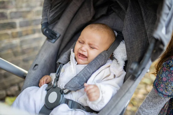 O bebê angustiado chora com os olhos fechados em um carrinho cinza ao lado de uma parede de tijolo — Fotografia de Stock