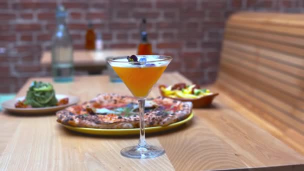 Cocktail op een houten tafel met frietjes, courgette noedels en een pizza erachter — Stockvideo
