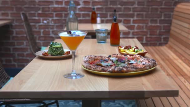 Cocktail on a wooden table with fries, zucchini noodles, and a pizza behind it — Stock Video