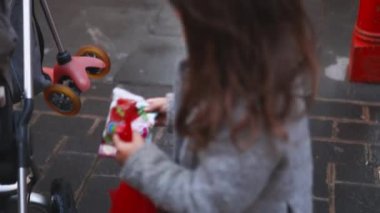 Küçük esmer kız gri bir bebek arabasının altına kırmızı plastik bir çanta koyuyor.