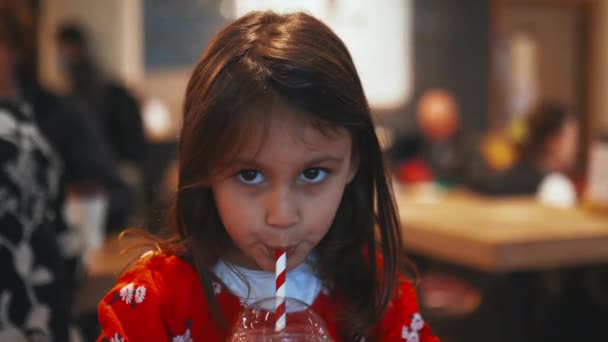 Little brunette girl sitting at a table and adorably sipping her red smoothie — Wideo stockowe