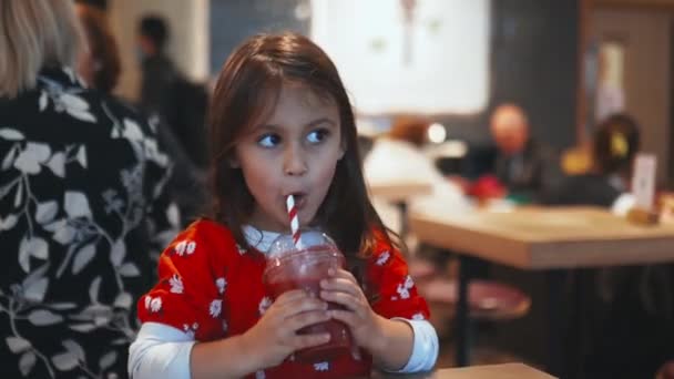 Egy kis barna lány ül az asztalnál, és édesen kortyolgatja a piros turmixát. — Stock videók