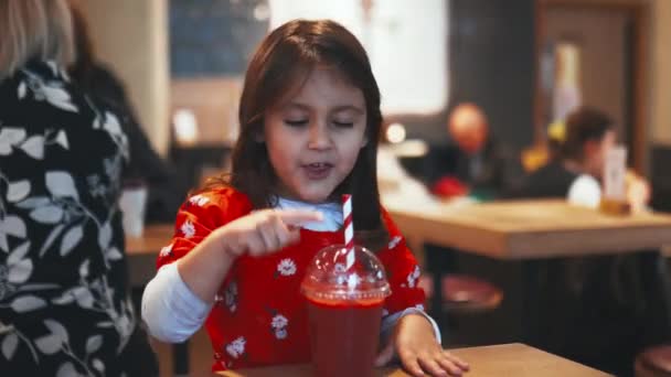 Little brunette girl sitting at a table and adorably sipping her red smoothie — Wideo stockowe