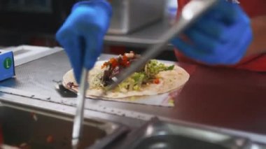 The hands of a man in blue gloves adding tomato slices to a meat burrito