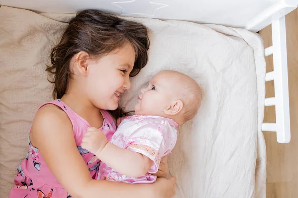 Joyful little girl lying down on a bed smiling at her baby sister and hugs her