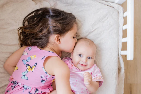 Little girl lovingly hugging and kissing her happy baby sister on a bed