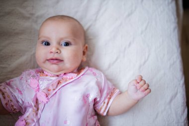 Asyalı pembe elbiseli meraklı ve tapılası bebek bir yatağa uzanıyor.