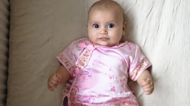 Glad och lekfull liten flicka i asiatisk rosa klädsel liggande på en säng — Stockvideo