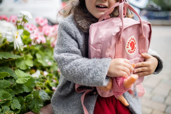 Маленькая девочка с цветами за спиной обнимает розовый рюкзак и маленькую куклу — стоковое фото