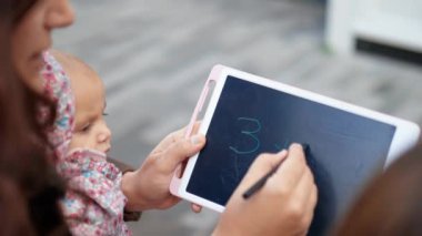Mutlu anne bebeğini kucağında tutuyor ve kızına tablette matematik öğretiyor.