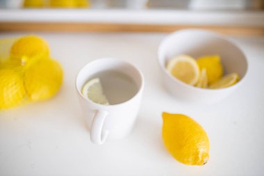 Limonlarla çevrili beyaz bir masada bir fincan limon çayı.