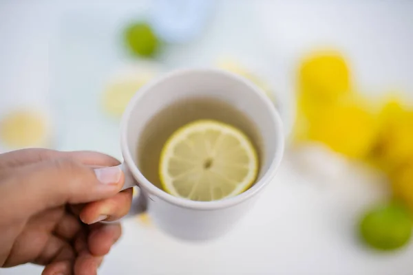 Mano femenina sosteniendo una taza de té de limón sobre limas y limones — Foto de Stock