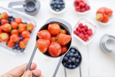 Kadın eli daha fazla çilek üzerinde ölçü kabı dolusu çilek tutuyor.