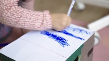 Küçük kız yoğun bir şekilde beyaz bir karton kutuyu mavi bir kalemle boyuyor.