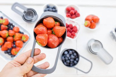 Kadın eli daha fazla çilek üzerinde ölçü kabı dolusu çilek tutuyor.