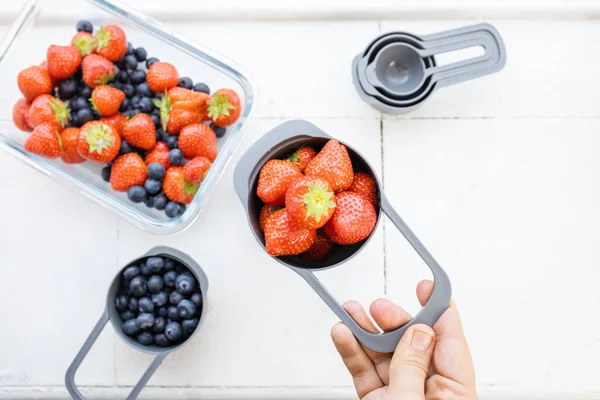 Kadın eli daha fazla çilek üzerinde ölçü kabı dolusu çilek tutuyor.