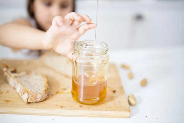 Niña recogiendo miel de un frasco junto a una rebanada de pan — Foto de Stock