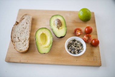 Dilimlenmiş ekmek, ikiye bölünmüş avokado, tahtanın üzerinde tohumlar ve domatesler.