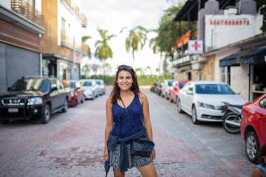 Güzel esmer kadın gülümsüyor ve tropik bir sokakta duruyor.