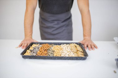 Siyah bir tepsi fındık ve tohumla beyaz bir masada oturan adam.